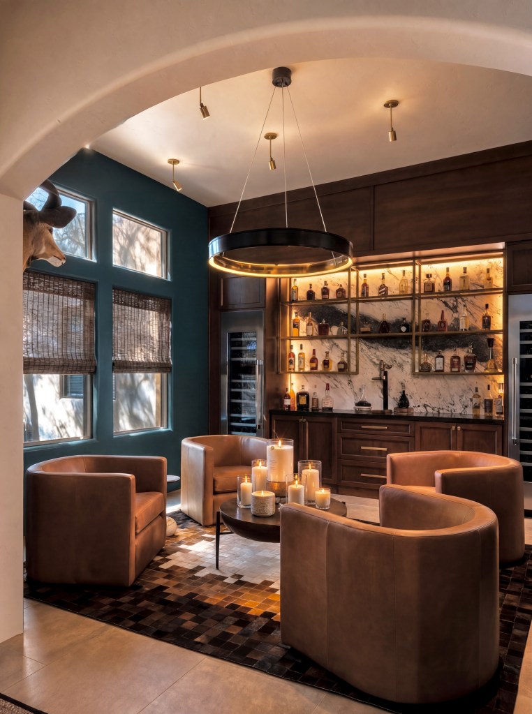 A stylish lounge area featuring four curved leather chairs arranged around a modern coffee table with candles. The backdrop includes a wooden bar with various bottles and a marble wall, complemented by large windows with blinds and a decorative wall mount.