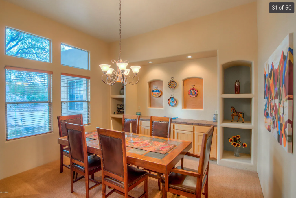 A dining room featuring a large wooden table surrounded by leather chairs, complemented by a chandelier. The walls are decorated with artwork and ceramic plates, and there's a display shelving unit with decorative items. Large windows provide natural light.